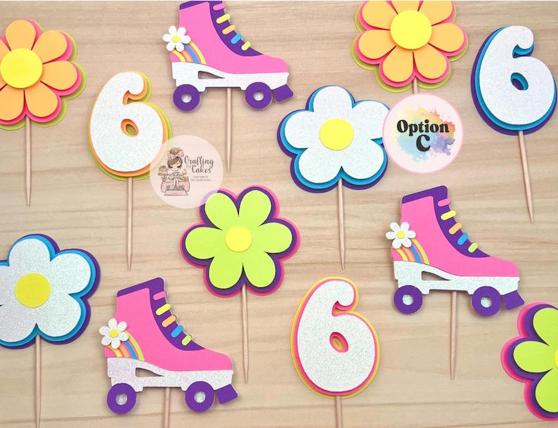 May include: A collection of colorful birthday cake toppers. The toppers include roller skates, flowers, and the number 6. The roller skates are pink with white accents and rainbow details. The flowers are yellow, pink, and blue.