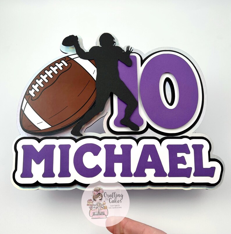 May include: A purple and white football-themed cake topper with the name "MICHAEL" and the number "10". The design includes a football player silhouette, a brown football, and a holographic border.