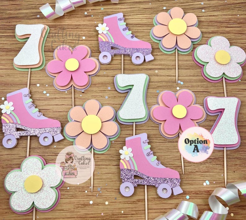 May include: A collection of decorative cupcake toppers featuring pink roller skates, flowers, and the number 7. The roller skates have glitter accents and small rainbows. The flowers are layered with pastel colors. The toppers are on wooden sticks.