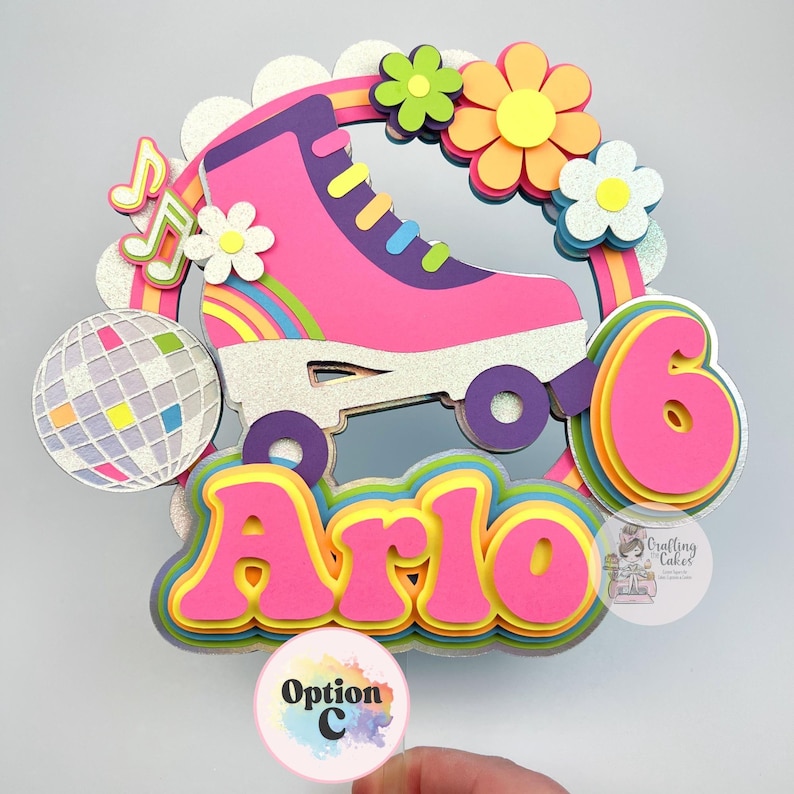 May include: A colorful cake topper featuring a pink roller skate, flowers, a disco ball, musical notes, and the name "Arlo" in layered letters. The number "6" is also included, suggesting a birthday theme.