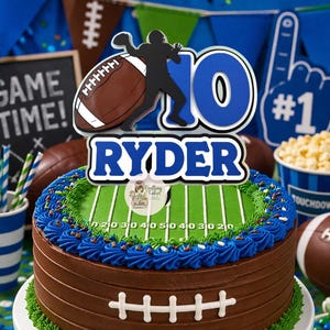 May include: A chocolate frosted cake decorated as a football field with blue frosting and a football-themed cake topper that says "RYDER" and "10". The cake is surrounded by football decorations and a "GAME TIME!" sign.