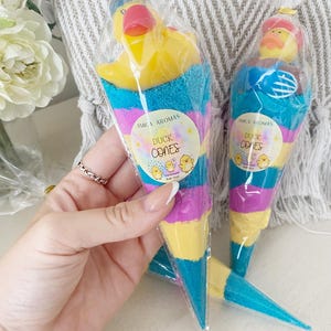 May include: Two cone-shaped bath dust products packaged in clear plastic. Each features a yellow rubber duck on top and layers of blue, purple, and yellow powder. A circular label reads Amica Aromas Duck Cones with small duck illustrations.