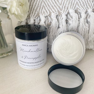 May include: A white jar with a black lid and a label that reads "AMICA AROMAS Marshmallow & Pineapple HANDMADE WHIPPED BODY BUTTER Shea Butter, Apricot Kernel Oil & Vitamin E"