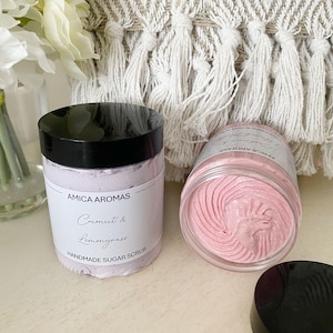 May include: Two jars of pink sugar scrub, one with a black lid and the other with a clear lid. The jar with the black lid has a white label that reads "Amica Aromas Coconut & Lemongrass Handmade Sugar Scrub".
