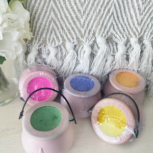 May include: Five small pink cauldron shaped bath bombs with a black handle. The bath bombs are filled with different coloured bath salts. The colours are pink, blue, orange, green, and yellow.