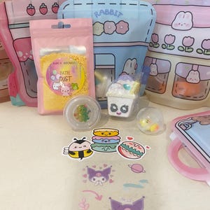 May include: A collection of items including three colourful plastic pouches with cartoon designs, a pink bag of yellow bath dust labelled Amica Aromas, a small clear container with a glittery bear, a dessert-shaped bath bomb, and various stickers.