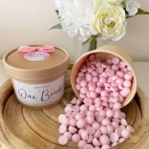 May include: Pink wax melts in the shape of beads in a brown paper container with a pink ribbon and the text "Wax Beads" and "Handmade Wax Melts".