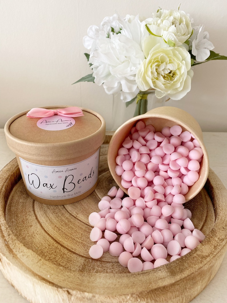 May include: Pink wax melts in the shape of beads in a brown paper container with a pink ribbon and the text "Wax Beads" and "Handmade Wax Melts".