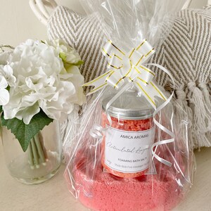 May include: A gift set with pink bath salts and a bath bomb, wrapped in cellophane and tied with a white and gold ribbon. The jar is labelled "Amica Aromas" and "Articulated Lingerie Foaming Bath Salts". The set is presented on a white surface.