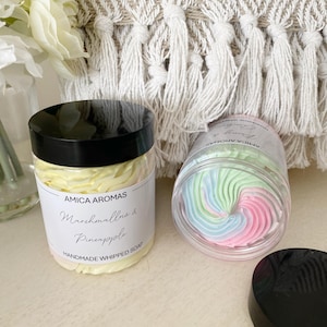 May include: Two jars of whipped soap. One jar is clear and shows a swirl of light blue, light green, and pink whipped soap. The other jar is white and has a label that reads "Amic Aromas Marshmallow & Pineapple Handmade Whipped Soap".