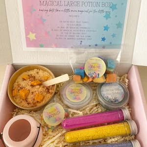 May include: A pink box filled with bath time treats for kids. The box contains a small pink cauldron, a tub of yellow bath dust, a bag of colourful bath sprinkles, a bag of crystal bath rocks, a jar of blue slime, a jar of white soap, and three test tubes filled with colourful bath dust. The box is labelled "Magical Large Potion Box" and "Making bath time a little more magical for the little ones."