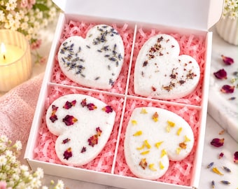 Puede incluir: Una caja de regalo blanca que contiene cuatro bombas de baño en forma de corazón sobre papel picado rosa. Cada bomba de baño está decorada con botánicos secos, incluyendo brotes de lavanda, pétalos de rosa y pétalos amarillos.