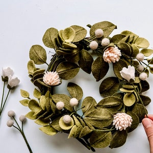 以下が含まれることがあります： オリーブグリーンの葉、薄いピンクの花、小さな白い実が付いた手作りのフェルトリース。追加のフェルトフラワーの茎がリースの左側に配置されています。リースは装飾品です。