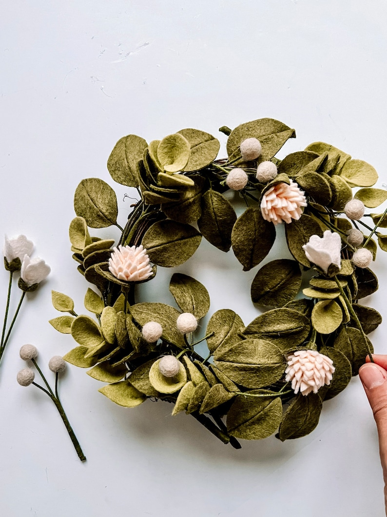 以下が含まれることがあります： オリーブグリーンの葉、薄いピンクの花、小さな白い実が付いた手作りのフェルトリース。追加のフェルトフラワーの茎がリースの左側に配置されています。リースは装飾品です。