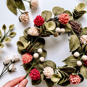 以下が含まれることがあります： 緑の葉、ピンクとクリーム色の花、白いベリーのアクセントが付いた、手作りのフェルトリース。背景には白黒のストライプのリボンが見えます。リースは装飾品です。