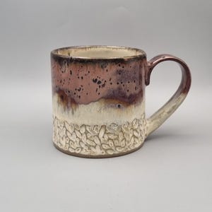 May include: A ceramic mug featuring a two-tone glaze with a dark, speckled mauve upper section and a cream-coloured lower section. The base of the mug is decorated with a raised, repeating leaf-like texture. It includes a curved handle on the side.