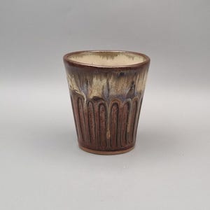May include: A ceramic tumbler featuring a tapered cylindrical shape with vertical fluted grooves. The exterior displays a deep reddish-brown glaze at the base that transitions into a creamy, dripped off-white glaze near the rim.