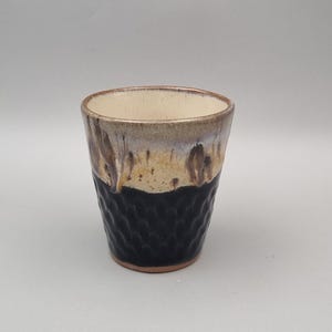 May include: A ceramic tumbler featuring a two-tone glaze with a cream-coloured upper section and a dark black lower section. The bottom half of the vessel has a textured, dimpled surface, while the interior is a smooth, light beige finish.