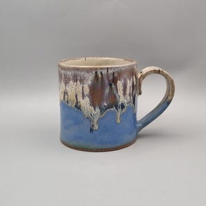 May include: A handcrafted ceramic mug. The base is blue, with a brown and cream-coloured top. It features a rounded handle and a textured, dripped glaze effect. Suitable for hot or cold drinks. The mug is approximately 10 cm tall.