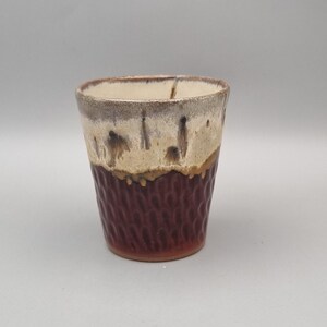 May include: A ceramic tumbler featuring a two-tone glaze design. The upper portion is a creamy off-white with dark brown drips, while the lower section is a deep reddish-brown with a textured, repeating vertical pattern across the surface.