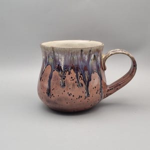 May include: A handcrafted ceramic mug with a distinctive glaze. The mug has a rounded body, a handle, and a speckled, reddish-brown base. The upper part has a purple and cream-coloured glaze, creating a layered effect. The interior is cream-coloured.