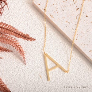 May include: A gold-toned necklace featuring a large, capital letter A pendant attached to a delicate chain. The letter A is positioned at an angle, and the text Paws & Knight is printed in the bottom right corner of the image.