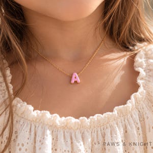 May include: A gold-toned chain necklace featuring a small, light pink letter A pendant. The pendant has a smooth, rounded finish and is centred on the delicate metal chain. The text Paws & Knight is visible in the bottom right corner.