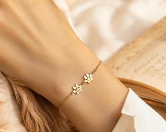 Könnte beinhalten: Ein zartes, goldfarbenes Kettenarmband mit zwei kleinen, polierten Pfotenabdrücken als Anhänger. Jeder Anhänger ist mit einem Namen graviert, was ein personalisiertes Schmuckstück mit einem schlichten, raffinierten Design aus Metallgliedern ergibt.