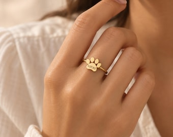 Könnte beinhalten: Ein goldfarbener Ring mit einem Pfotenabdruck-Design auf der Vorderseite. In die Mitte des Pfotenabdrucks ist der Name LUCAS in Großbuchstaben eingraviert. Der Ring hat eine dünne, glatte Ringschiene, die den Finger umschließt.