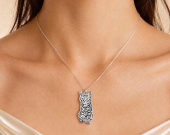Puede incluir: Un collar de tono plateado con un colgante grabado y detallado en forma de gato atigrado sentado. El colgante cuelga de una cadena fina y delicada de color plateado que descansa sobre la clavícula, mostrando un retrato felino realista.