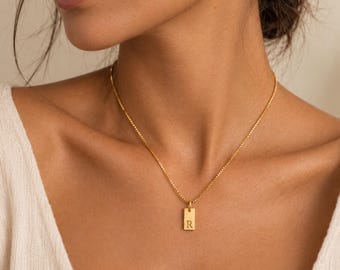 Könnte beinhalten: Eine goldfarbene Halskette mit einer feinen Boxkette und einem kleinen, rechteckigen Anhänger. Der Anhänger ist mit dem Großbuchstaben R in einer Serifenschrift graviert. Die Kette liegt am Hals an und hat ein poliertes, metallisches Finish.