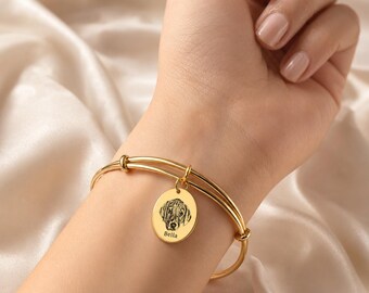 Könnte beinhalten: Ein goldfarbenes Armband mit einem runden Anhänger, der eine detaillierte Strichzeichnung eines Hundegesichts und den darunter eingravierten Namen Bella zeigt. Das Armband besteht aus zwei dünnen, glatten Metallreifen, die am Anhänger zusammenlaufen.