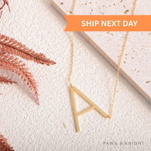 May include: A gold-toned necklace featuring a large, capital letter A pendant suspended from a delicate chain. The pendant is attached to the chain at two points, creating a side-oriented look. The text SHIP NEXT DAY is visible.