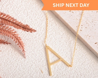 Puede incluir: Un collar de tono dorado con un colgante grande en forma de letra A mayúscula suspendido de una cadena fina. El colgante está unido a la cadena en dos puntos, creando un aspecto lateral. El texto SHIP NEXT DAY es visible.