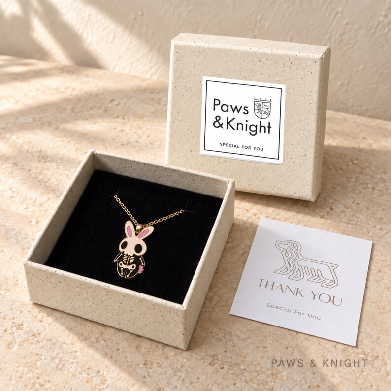 Puede incluir: Un collar dorado con un colgante en forma de conejo esqueleto con orejas rosas, presentado en una caja beige moteada. La tapa tiene una etiqueta con el texto Paws & Knight Special For You y una tarjeta con una ilustraci&oacute;n de un perro.