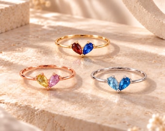 Könnte beinhalten: Drei dünne Metallringe, jeweils mit zwei tropfenförmigen Edelsteinen in Herzform. Ein goldener Ring mit roten und blauen Steinen, ein roségoldener Ring mit gelben und rosa Steinen und ein silberner Ring mit hell- und dunkelblauen Steinen.