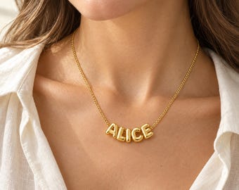 Könnte beinhalten: Eine goldfarbene Halskette mit einer feinen Kette und fünf einzelnen, abgerundeten Buchstabenanhängern im Ballon-Stil, die den Namen ALICE bilden. Die metallischen Buchstaben haben eine glatte, reflektierende Oberfläche und sind mittig angeordnet.