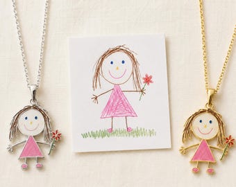 Könnte beinhalten: Zwei Halsketten mit Anhängern in Form einer Kinderzeichnung einer Person in einem rosa Kleid, die eine rote Blume hält. Eine Kette ist silberfarben, die andere goldfarben. Eine quadratische Karte zeigt die Originalzeichnung der Figur.