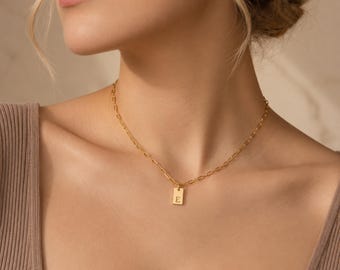 Könnte beinhalten: Eine goldfarbene Halskette mit einer zarten Kette und einem kleinen, rechteckigen Anhänger, der mit dem Buchstaben E graviert ist. Der Anhänger hängt mittig an der Kette, die am Schlüsselbein aufliegt und ein poliertes Metallfinish aufweist.