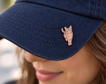 Könnte beinhalten: Eine marineblaue Baseballkappe mit einer kleinen, hellbraunen Holzanstecknadel in Katzenform am Schirm. Die Katzenbrosche zeigt detaillierte, eingravierte Linien, die das Fell, die Ohren und den Schwanz darstellen. Die Kappe hat sichtbare Nähte.