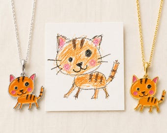 Könnte beinhalten: Zwei Halsketten mit orangefarbenen Katzenanhängern mit schwarzen Streifen und rosa Wangen. Ein Anhänger ist silberfarben, der andere goldfarben. Dazwischen liegt ein quadratisches Papier mit einer Buntstiftzeichnung einer Katze. Text: PAWS & KNIGHT.