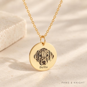 Puede incluir: Un collar con colgante circular dorado que presenta el retrato grabado de la cara de un perro y el nombre Bella inscrito debajo de la imagen. El colgante cuelga de una delicada cadena de eslabones dorada.
