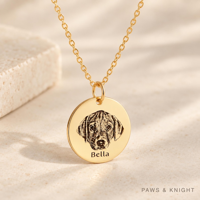 Puede incluir: Un collar con colgante circular dorado que presenta el retrato grabado de la cara de un perro y el nombre Bella inscrito debajo de la imagen. El colgante cuelga de una delicada cadena de eslabones dorada.