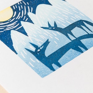 Peut inclure: Une estampe linogravure bleue et blanche représentant deux loups stylisés hurlant à la lune dans un paysage montagneux enneigé.