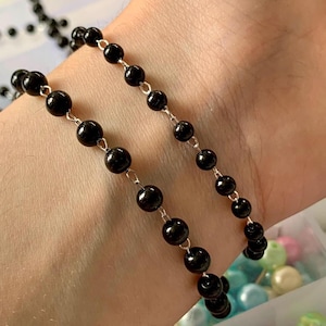 May include: Two black beaded bracelets with silver-colored chain links are displayed on a wrist. The beads are round and shiny. The bracelets are next to a partially visible strand of similar beads.