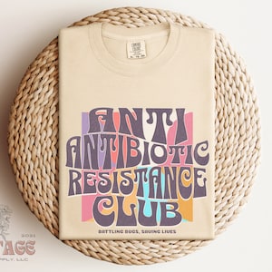 May include: A beige t-shirt with a retro-style graphic that says "Antibiotic Resistance Club" in colorful, wavy letters. The text is surrounded by a pink, orange, and blue background. The bottom of the shirt says "Battling Bugs, Saving Lives".