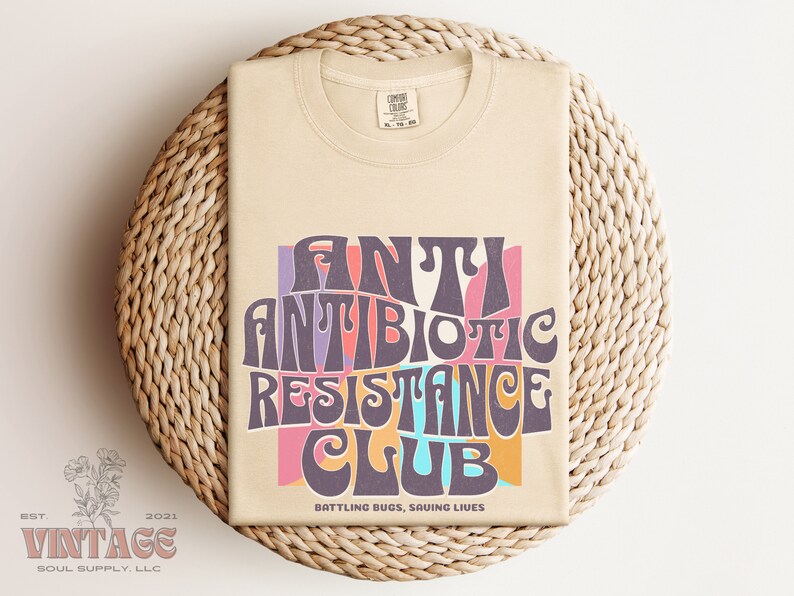 May include: A beige t-shirt with a retro-style graphic that says "Antibiotic Resistance Club" in colorful, wavy letters. The text is surrounded by a pink, orange, and blue background. The bottom of the shirt says "Battling Bugs, Saving Lives".