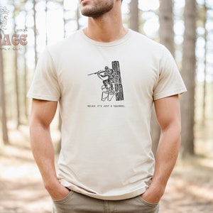 May include: A beige t-shirt with a black graphic of a person climbing a tree with the text "Relax, it's just a squirrel." printed below the graphic.