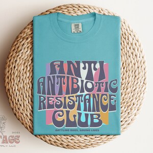 May include: A light blue t-shirt with a retro-style graphic that says "Antibiotic Resistance Club" in colorful letters. The graphic is surrounded by a pink, yellow, and blue background. The text below the graphic says "Battling Bugs, Saving Lives".