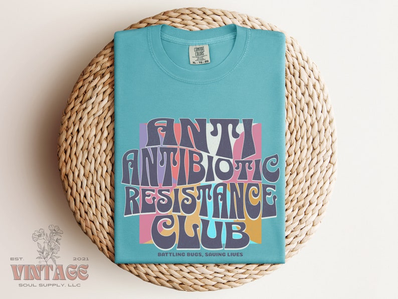 May include: A light blue t-shirt with a retro-style graphic that says "Antibiotic Resistance Club" in colorful letters. The graphic is surrounded by a pink, yellow, and blue background. The text below the graphic says "Battling Bugs, Saving Lives".